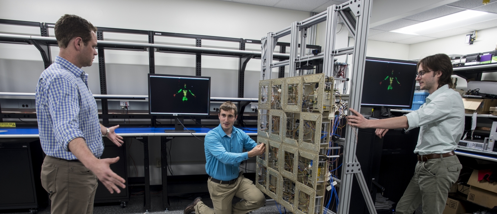 Researchers test the prototype standoff microwave imaging system. The antennas emit radio signals that reflect off the person standing in front of the array; the system processes the reflections to create the image on the monitors in the background.