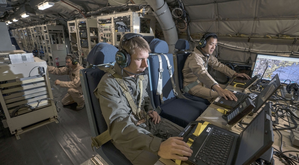 Two personnel are seated inside a military test aircraft operating communications equipment.