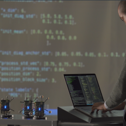 A researcher stands on a laptop. In front of him are a few small radios, and a large projector screen showing data.